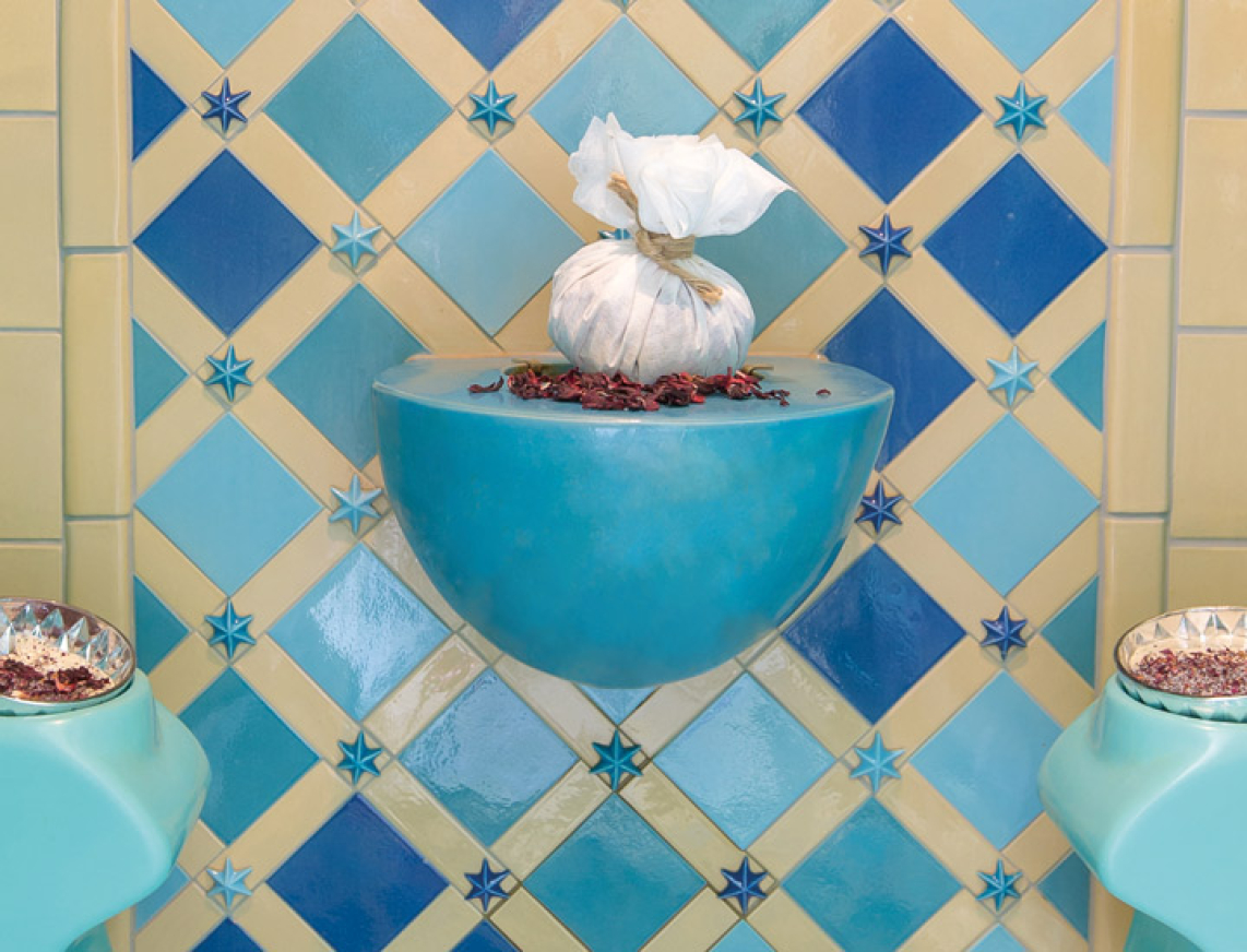 Blick auf einen stilvoll gestalteten orientalischen Rhasul mit türkisfarbenen Sitzflächen und einer kunstvoll gefliesten Wand in Blau- und Beigetönen. In der Mitte befindet sich ein dekorativer Dampfbrunnen.