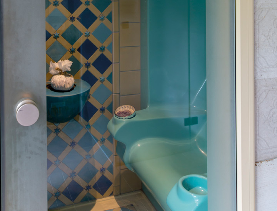 Blick auf einen stilvoll gestalteten orientalischen Rhasul mit türkisfarbenen Sitzflächen und einer kunstvoll gefliesten Wand in Blau- und Beigetönen. In der Mitte befindet sich ein dekorativer Dampfbrunnen.