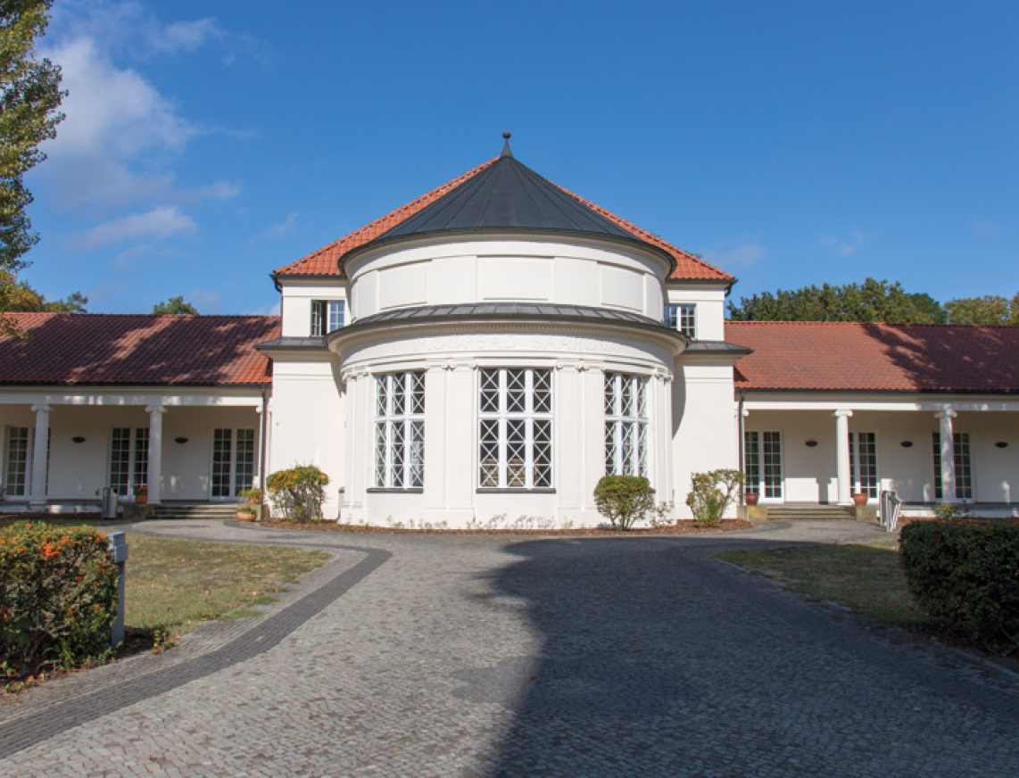 Das Alte Moorbad Bad Saarows. Weißes Gebäude mit runder Kuppelform an der Vorderseite des Gebäudes. Weiße Hauswände, weiße Säulen und große französische Fenster geben dem Gebäude sein historisches Flair. Ein gepflasteter Weg führt von der Straße bis zum Eingang des Moorbades.