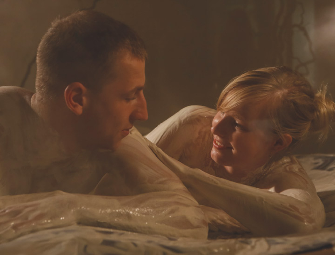 Junges Pärchen cremt sich gegenseitig mit einem weißen Cremepeeling auf der Membranliege des Salve-in-terra der SaarowTherme ein. Lächelnd genießen Sie diesen sinnlichen Moment der Zweisamkeit.