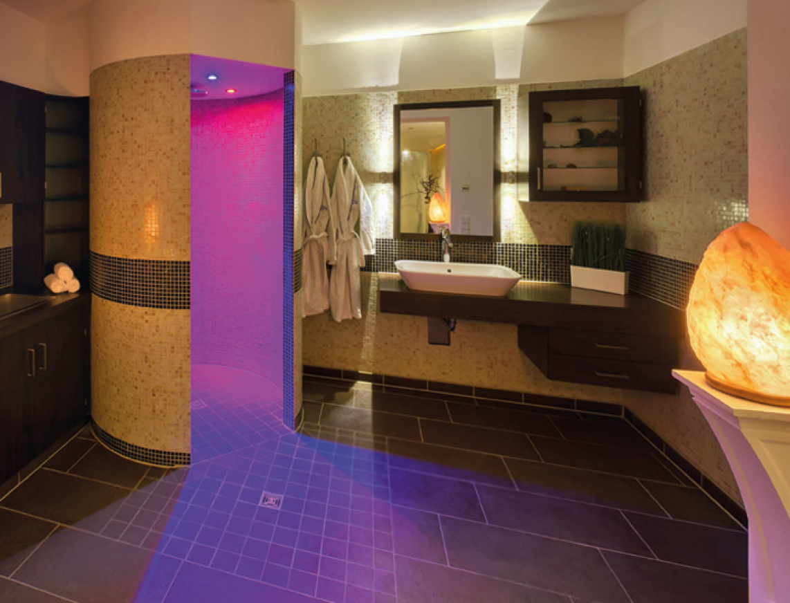 Ansicht des Nebenbereiches der Salve in terra. Eine Runde Dusche mit Farblicht, großes Waschbecken mit Ablageflächen und Garderobehaken mit zwei Bademäntel sind zu sehen. Im Vordergrund schafft ein beleuchteter Sandstein eine angenehme Atmosphäre im Raum.
