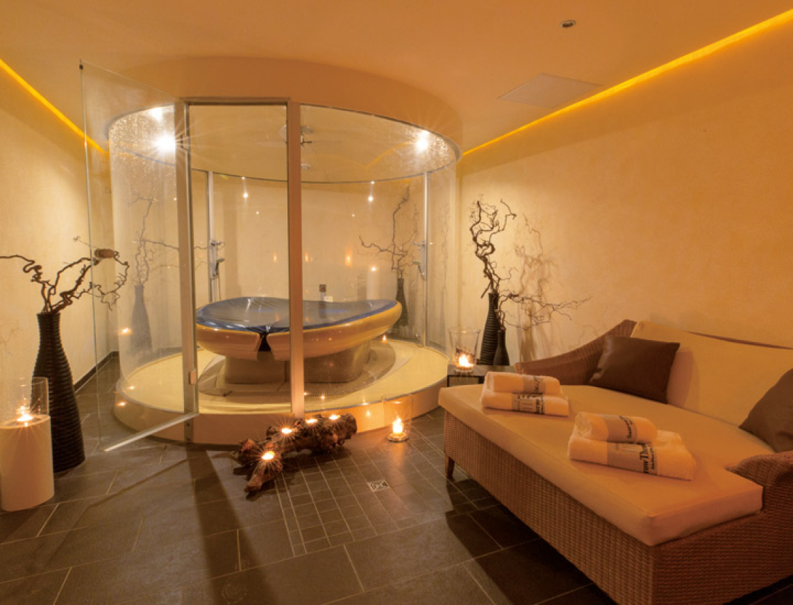 Ein stilvoll beleuchteter Wellnessraum mit einer modernen Membranliege in einer gläsernen Kabine. Umgeben von dekorativen Elementen wie Ästen und Kerzenlicht entsteht eine warme und entspannte Atmosphäre. Daneben liegt ein Handtuch auf einer bequemen Duoliege.
