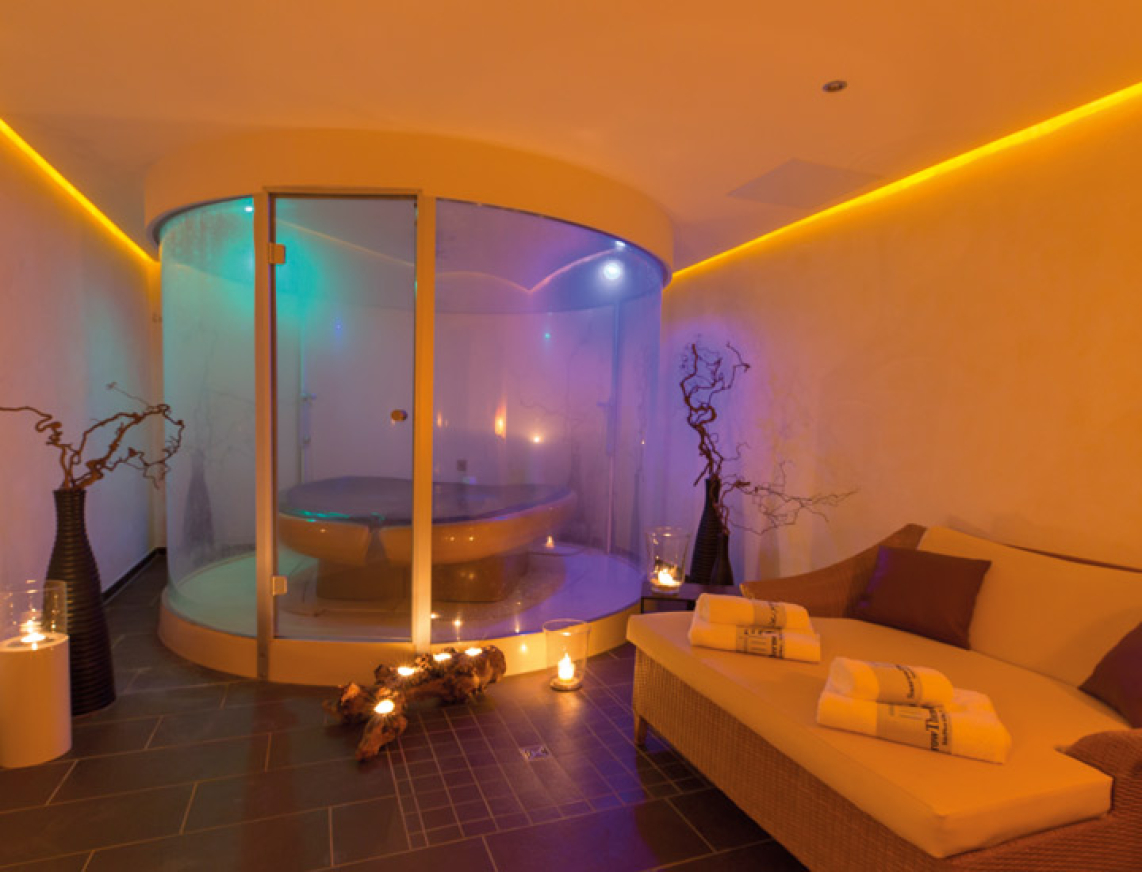 Das Salve-in-terra der SaarowTherme. Ein großer Raum mit in der Mitte eine Membranliege von einer Glaswand umgeben und in einem bläulichen Licht getaucht. Eine Duoliege mit Liegefläche zum Verweilen und Kerzenschein im Raum vermitteln eine romantische Atmosphäre.
