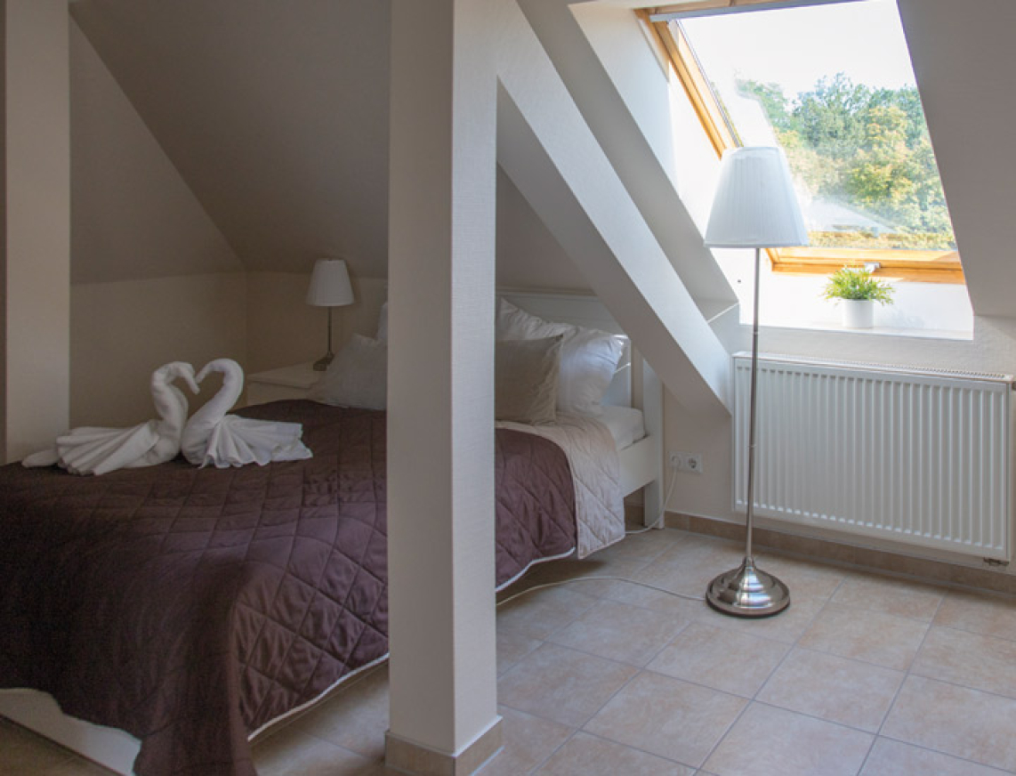 Schlafbereich im Apartment im Alten Moorbad des SaarowCentrums. Ein großes Dachfenster wirft ein angenehmes Tageslicht auf dem Doppelbett, dass neben dem Fenster steht. Das Bett ist mit einer braunen Tagesdecke bezogen und am Bett steht ein kleiner Beistelltisch mit einer weißen Stehlampe.