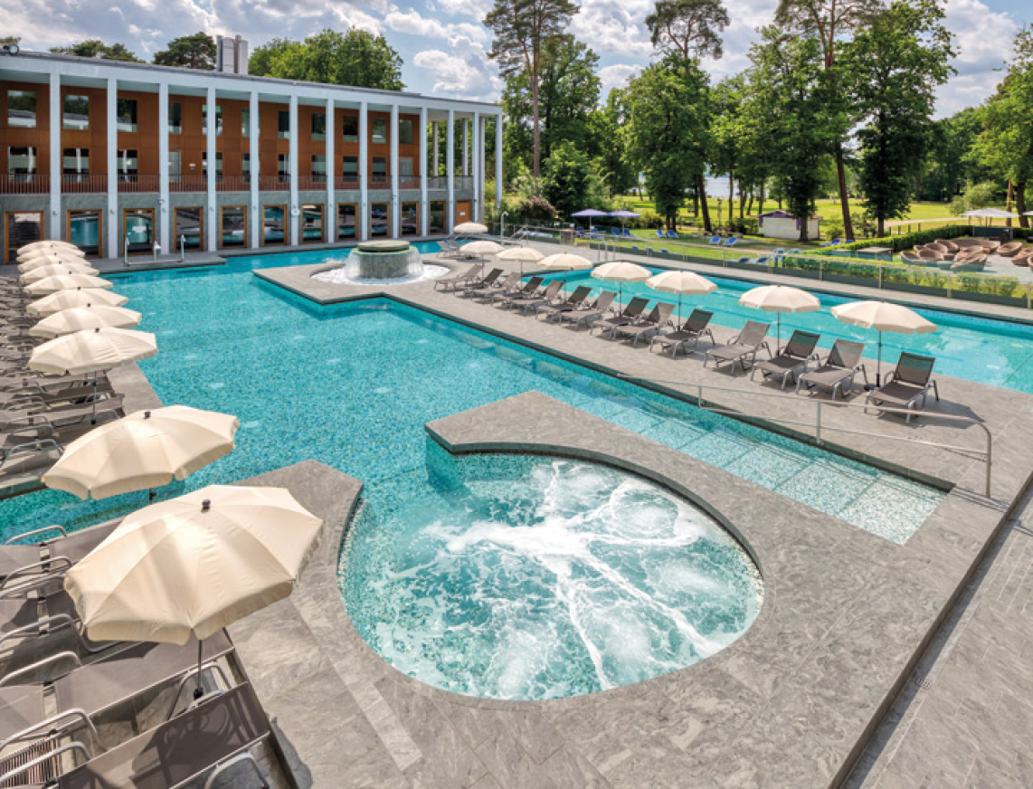Großes Thermalbad im Außenbereich mit Whirlpool, Sonnenliegen, Strömungskanal und weitläufigem Sportbecken zum Erholen und Entspannen. Im Hintergrund ist der Kurpark Bad Saarow mit seiner grünen Natur zu sehen.