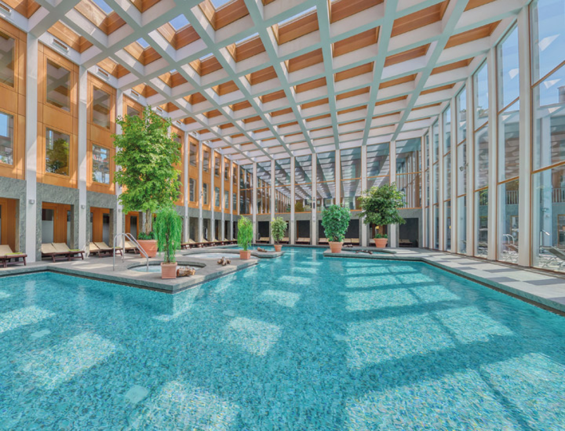 Lichtdurchflutetes Thermalbad mit großen Fenstern und Deckenkuppel. Türkisblaues Wasser und grüne Pflanzen vermitteln ein Ort zum Entspannen und Erholen.