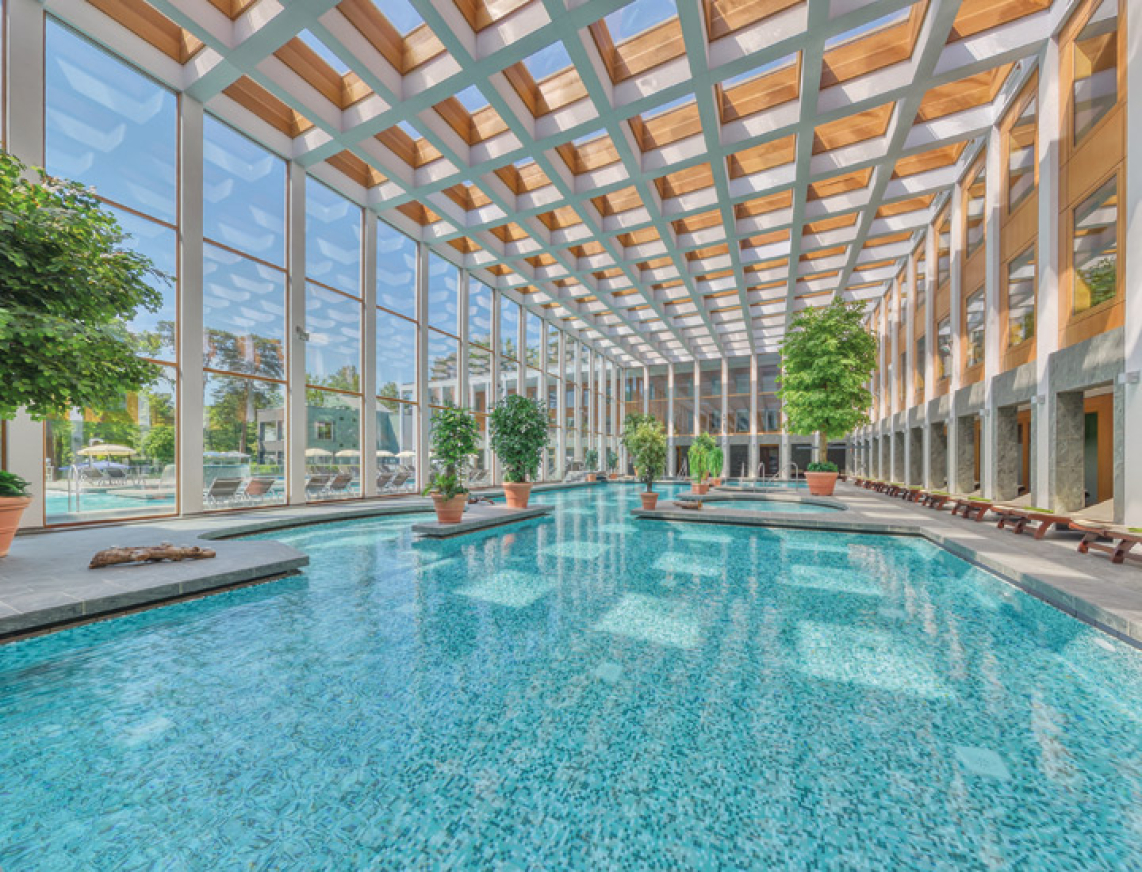 Lichtdurchflutetes Thermalbad mit großen Fenstern und Deckenkuppel. Türkisblaues Wasser und grüne Pflanzen vermitteln ein Ort zum Entspannen und Erholen.