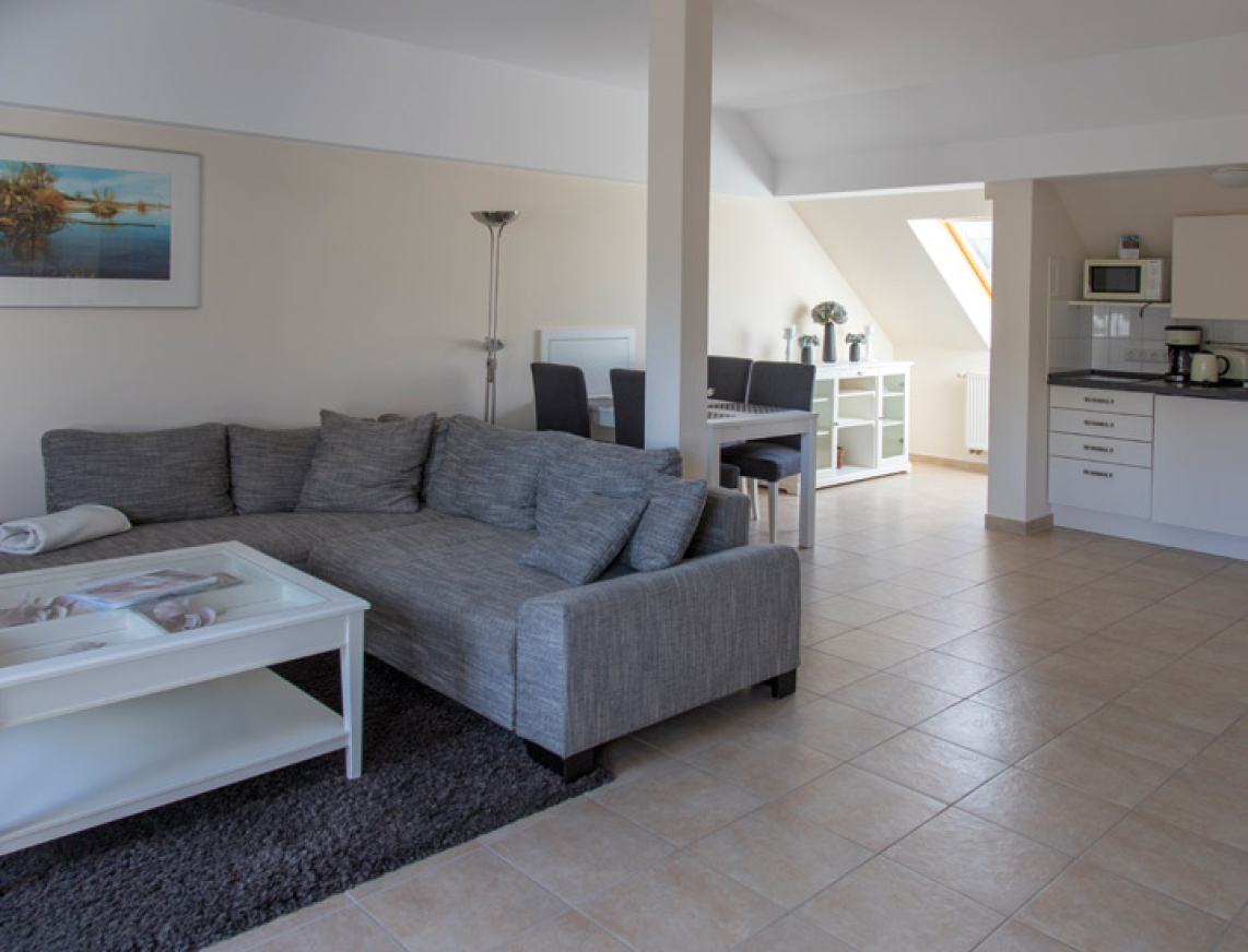 Helles Wohnzimmer mit grauem Ecksofa, weißem Couchtisch und offener Einbauküche. Im Hintergrund ein Essbereich unter einem Dachfenster.