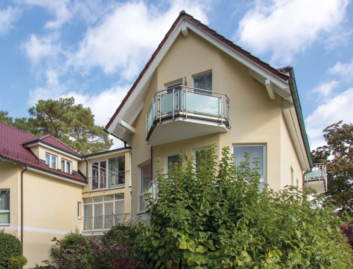 Zweistöckiges hellgelbes Wohnhaus mit roten Dächern und kleinen Balkonen, umgeben von Grünpflanzen und Bäumen unter blauem Himmel.