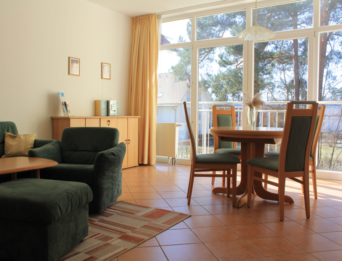 Helles Wohnzimmer mit großen Fenstern und Zugang zum Balkon, ausgestattet mit grünem Sofa, Sessel, Esstisch und Holzkommode.