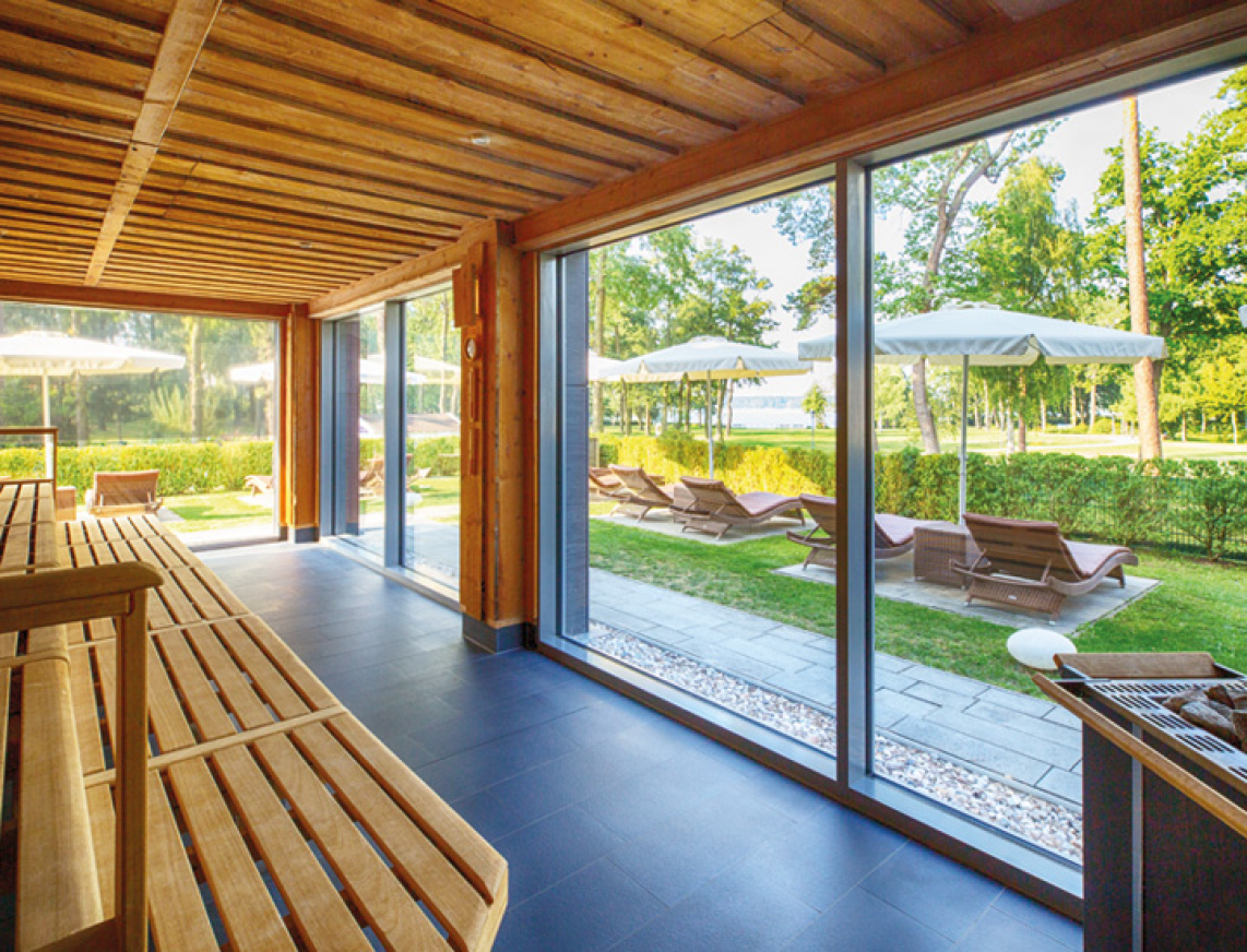 Innenansicht der großzügigen, lichtdurchfluteten KeloSauna mit Holzverkleidung und Panoramafenster. Draußen sind Liegestühle und Sonnenschirme im gepflegten Gartenbereich zu sehen.