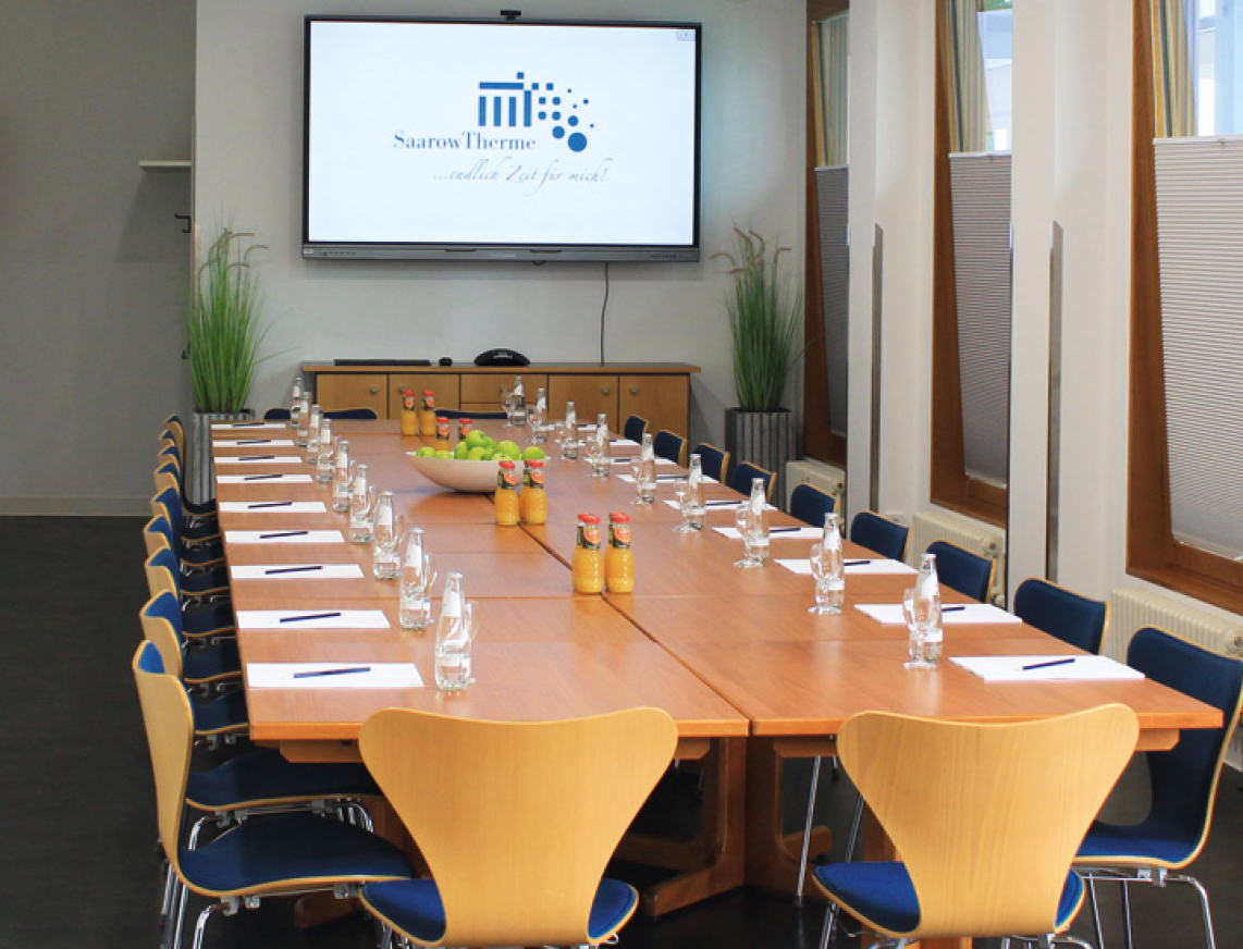 Tagungsraum in der SaarowTherme mit langem Tisch, blauen Stühlen und großem Bildschirm mit Logo. Auf dem Tisch stehen Wasserflaschen, Orangensaft, Schreibblöcke und Bleistifte – ideal für Seminare, Workshops und Tagungen in Bad Saarow.