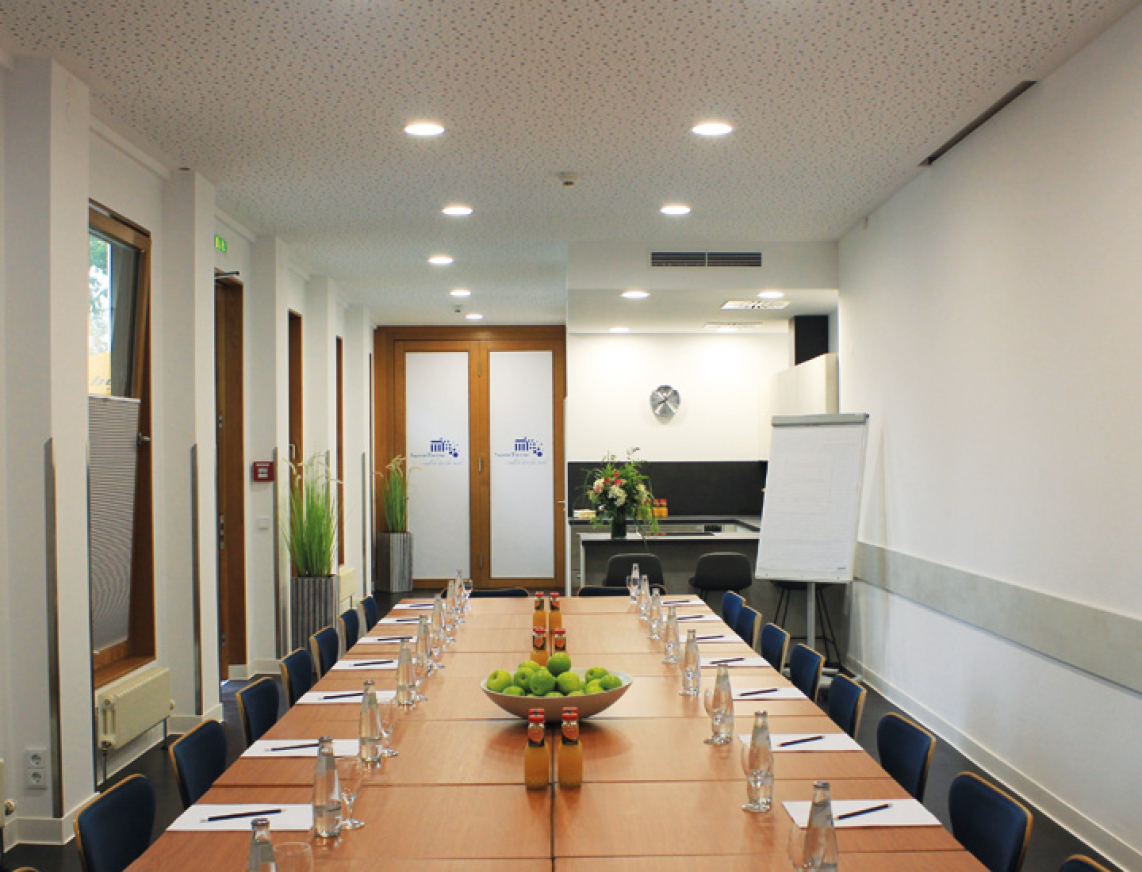 Tagungsraum mit langem Tisch, blauen Stühlen und großem Bildschirm mit dem Logo der SaarowTherme. Auf dem Tisch stehen Wasserflaschen, Orangensaft, Schreibblöcke und Bleistifte, vorbereitet für eine Tagung. Im Hintergrund ist die integrierte Küche des Tagungsraums zu sehen.