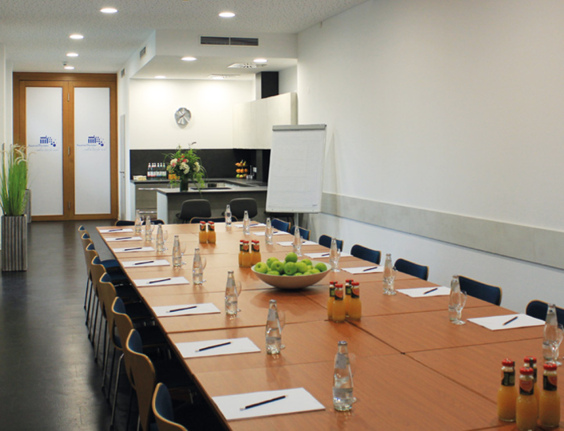 Tagungsraum in der SaarowTherme mit langem Konferenztisch, ausgestattet mit Schreibblöcken, Bleistiften und Getränken. Im hinteren Bereich befindet sich eine voll ausgestattete Küche – ideal für Seminare, Tagungen und Workshops in Bad Saarow.