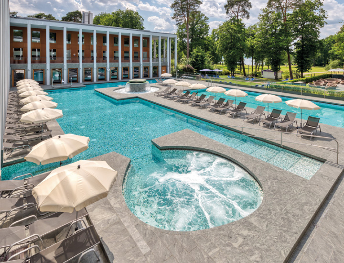 Außenbereich der SaarowTherme in Bad Saarow mit Thermalsolebecken, großem Whirlpool sowie Liegestühlen und weißen Sonnenschirmen am Beckenrand. Im Hintergrund liegt der grüne Kurpark direkt am Scharmützelsee.