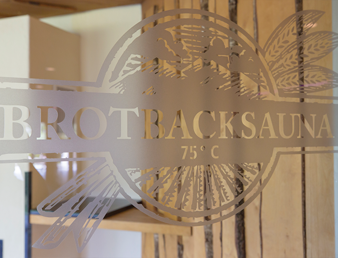 Glaswand mit „BROTRACKSAUNA 75°“-Logo, dahinter Holzsauna und Brotbackofen